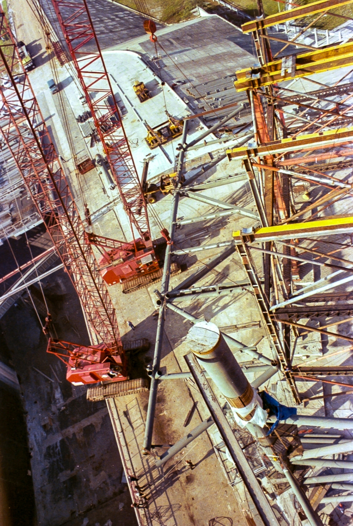Large-format photograph: With the crawler tracks of one crane literally hanging over the side of the Flame Trench West Wall, Wilhoit Steel Erectors begins carefully lifting the RSS Bottom Truss Weldment. The truss is 160 feet long, weighs over 150,000 pounds, and will be placed on top of the Falsework which extends out of frame to the right. In this image, the Truss has traveled roughly 30 feet upwards, in a horizontal orientation, and has roughly another 50 vertical feet to go, before Wilhoit can then begin the ticklish processes required to get it properly out and across the full width of the Falsework to its final resting place. Photo courtesy Eugene Hajdaj.